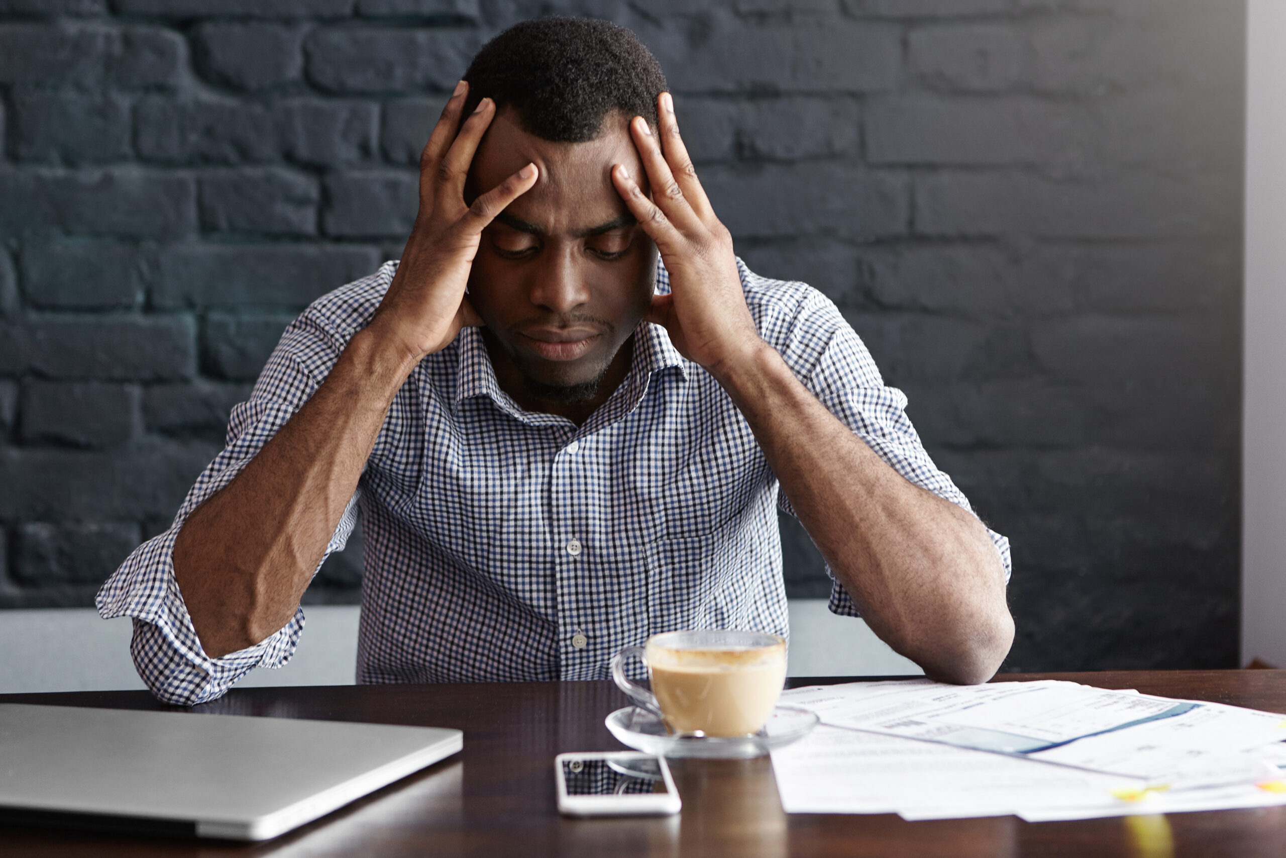 Frustrated young African-American businessman having bad headache, squeezing his temples, feeling stressed at work, sitting at desk with generic laptop computer, documents, mug and mobile phone